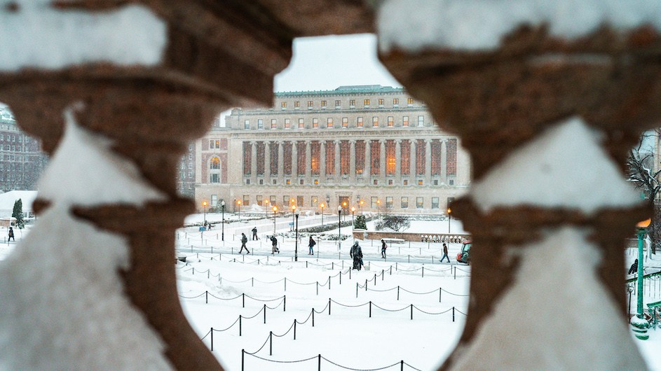 A photo of shot through a marble bannister looking at snowy scene with people walking and a large white marble building with columns in the background