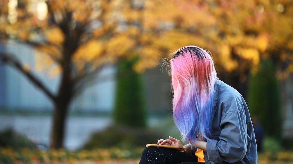 A photo of a young woman with pink, purple and white hair sitting on the ground with fall foliage in the background.