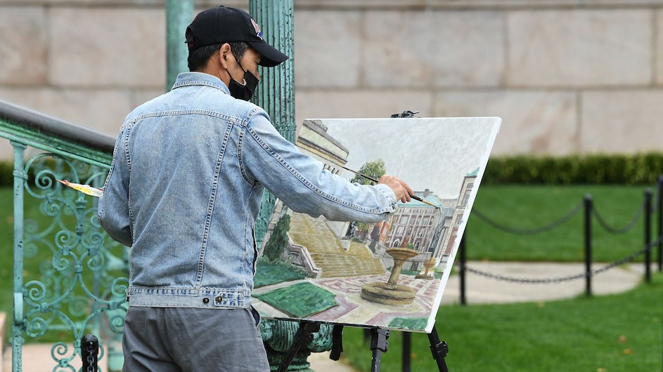 A man with a baseball cap and a mask painting on an easel outside near a staircase.