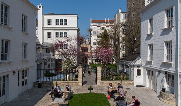A sunlit courtyard with students at tables, surrounded by white buildings and trees