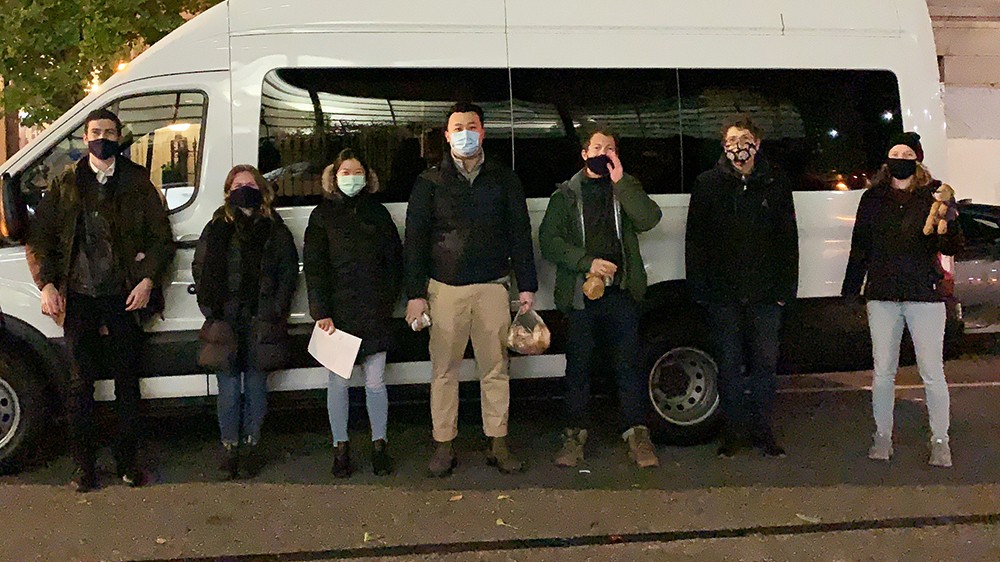 A photo of seven people wearing masks standing in front a white van on the street at night.