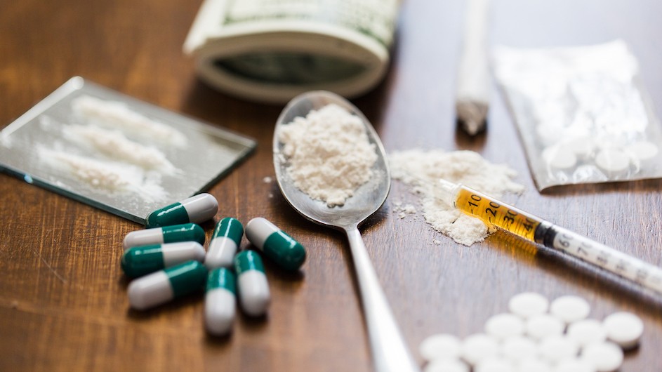 Green and white pills, a spoon with white powder, a syringe, money and lots of pills on a table