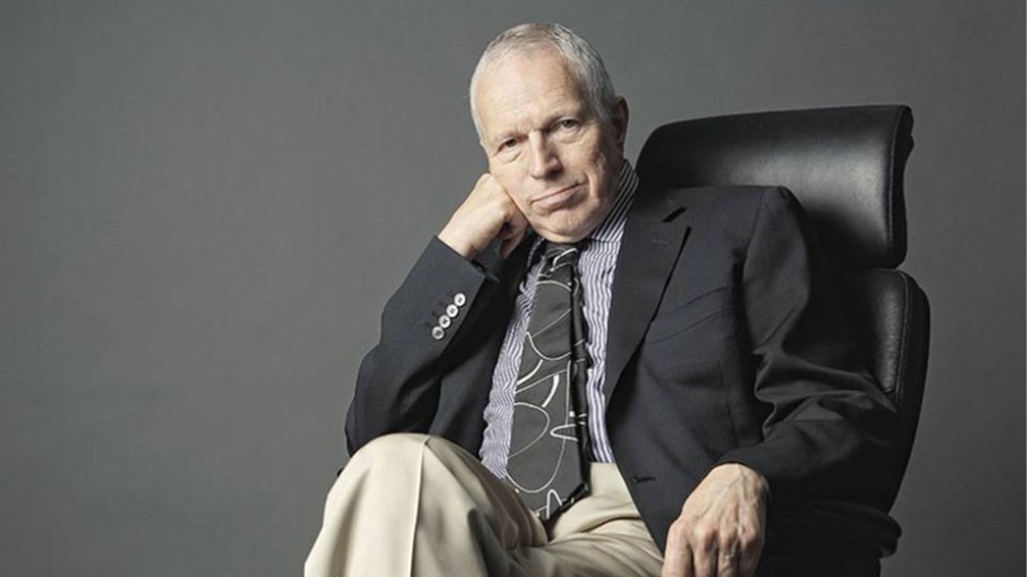 An image of a man wearing a tie and jacket seated in a chair slightly resting his head on his hand.