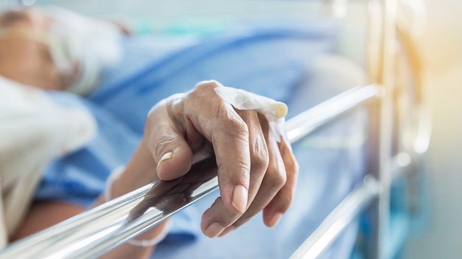 Close up of an elderly patient's hand with intravenous catheter for injection plug lying in a hospital ward room bed.