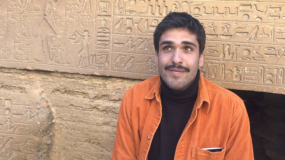GSAPP graduate Ibrahim Kombarji on a recent trip to Egypt. Ibrahim Kombarji: A young man in an orange shirt in front of a wall with carvings.