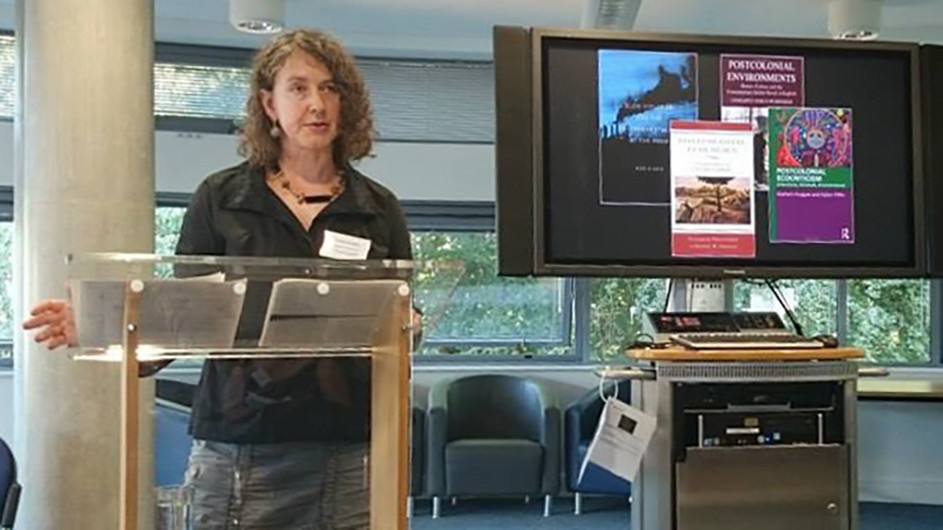Professor Jennifer Wenzel has written a new book, 'The Disposition of Nature: Environmental Crisis and World Literature.' Woman with light brown curly hair standing at a podium with a screen in the background highlighting a talk she is giving