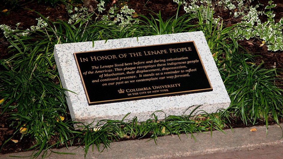 A black plaque with gold writing and gold trim on a stone displayed in a garden at Columbia University honoring the Lenape People
