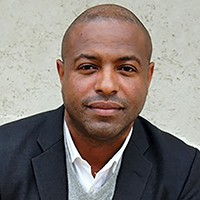 A man in a dark jacket and white shirt in front of a white-gray wall.