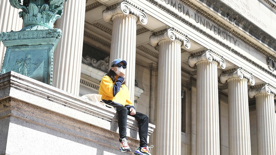 A student sits in front of the column with a yellow sweatshirt, baseball cap, sunglasses, and mask on.on. 