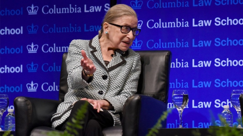 Justice Ruth Bader Ginsburg is seated in a black leather chair against a Columbia Law School backdrop, gesturing to a crowd of onlookers. 