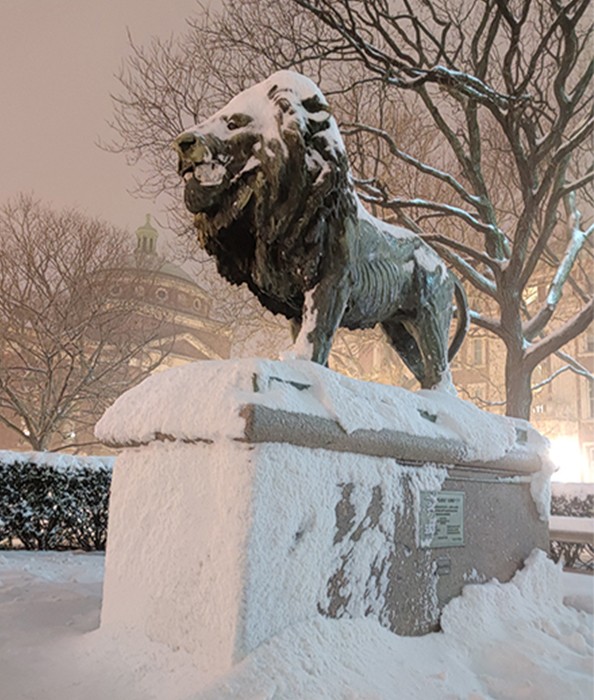 A grey statue of a lion on a stone pedestal is enshrouded in a blanket of snow. 