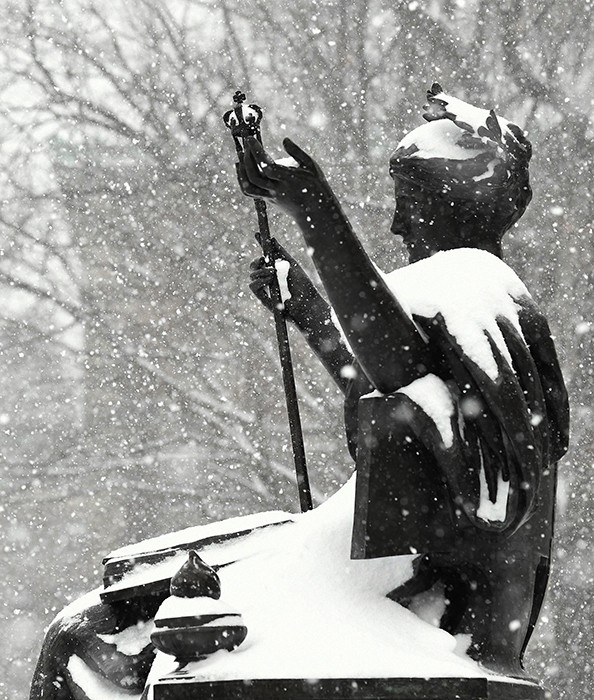 A black statue of Columbia's Alma Mater, with hands and scepter upraised, is enshrouded in a blanket of snow as snowflakes swirl around her.