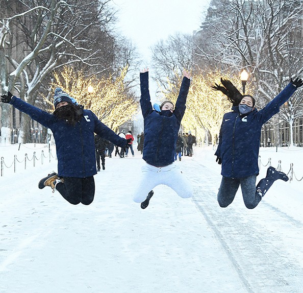 Three students in masks, hats, gloves, and jackets, jump on College Walk sidewalk in front of trees covered in snow with holiday lights.