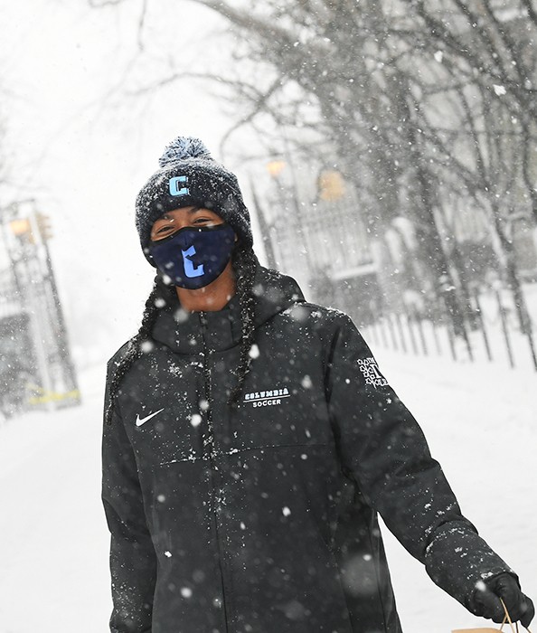A woman in a black Ivy League jacket and Columbia-emblazoned hat and mask walks on a snowy sidewalk.
