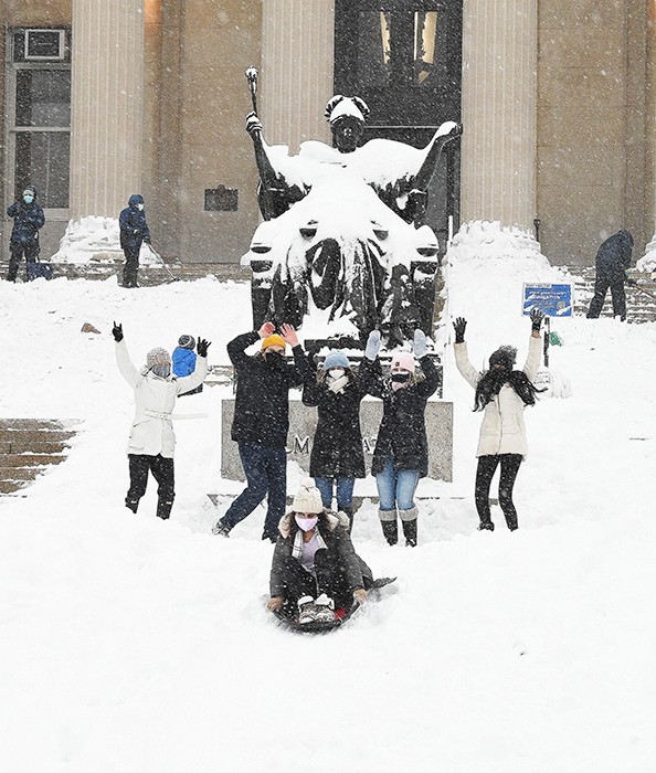 A group of students in masks and hats stand with hands raised in front of the Alma Mater statue, covered in snow, as another student sleds down Low Steps.