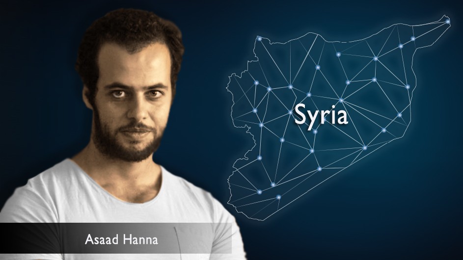 A man with short, dark hair, a dark mustache and beard, in a white t-shirt, against a map of syria drawn out in stars.