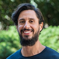 Headshot of a man with short, dark hair, a dark mustache and a dark beard, in a dark blue top