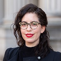 Headshot of a woman with medium-length dark hair and glasses, in a dark top and coat