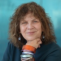 A headshot of a woman with brown shoulder length hair and she's wearing many bracelets and a blue shirt