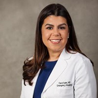 A woman with long brown hair and in a white lab coat and blue dress poses against a grey background.