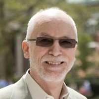 A close-up photo of a man in sunglasses standing against a leafy background with a white beard and white hair.