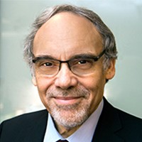 A man with glasses, grey hair, and a beard poses in a black suit jacket against a grey background.