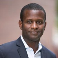 A black man with short black hair poses in a white shirt and blue suit jacket against a neutral background.