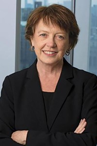 A woman with short, medium brown hair, in a black suit standing in front of a window