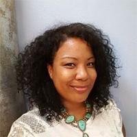 A headshot of a woman with shoulder length dark curly hair who is wearing a cream colored shirt and a green necklace