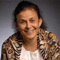 A tan woman with black hair is photographed from the shoulders up in a tan pashmina and white shirt against a grey background.