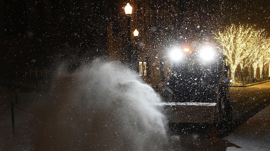 A snow plow moving snow at night with lit trees in the background