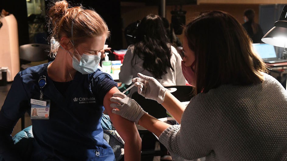 A woman, with blonde hair tied in a bun wearing a mask and blue scrubs, is getting the SARS-CoV2 vaccine to protect against COVID-19.