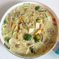 Carrots, baby corn, tofu, and broccoli sit in a white pot of yellow-green sauce. 