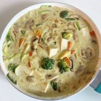 Carrots, baby corn, tofu, and broccoli sit in a white pot of yellow-green sauce.