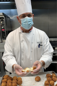 Chef Mike of Columbia University: A man in a mask, white chef's hat and chef shirt with a mask holding a cheesy rice ball in his outstretched gloved hands.