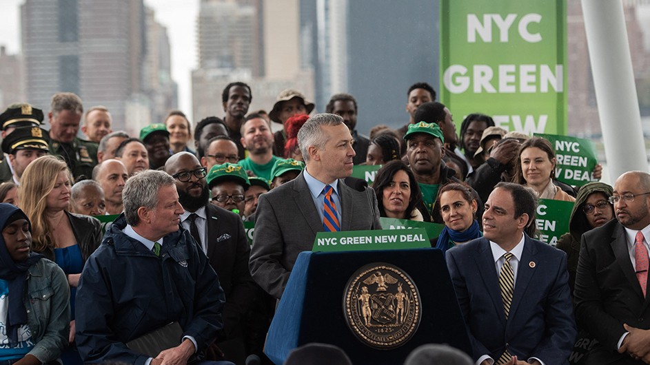 A man standing at a podium in front of a crowd, surrounded by banners titled, "NYC Green New Deal."