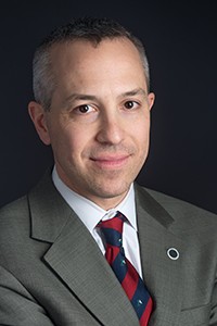 Headshot of a man with short hair in a grey suit, white shirt, and red and blue tie.