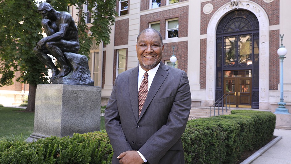 Michael DiVito A man in a grey suit, standing outside in front of a brick building and a statue of a man sitting, thinking, resting his head on his hand.