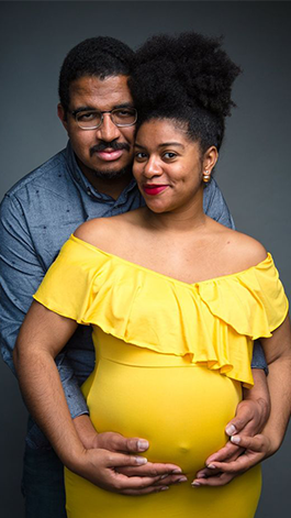 A husband in a blue shirt hugs his pregnant wife in a yellow dress from behind.