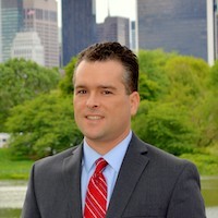 Jeff Schlegelmilch: A headshot of a man with short dark hair who is wearing a grey suit, light blue shirt and a red tie. Trees and buildings are in the background.