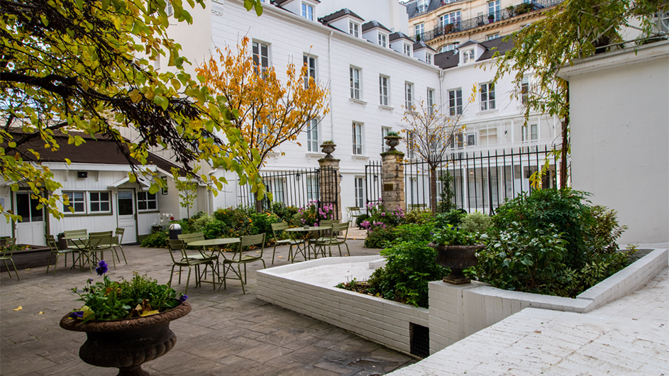 A sunlit courtyard with trees, plants, and tables, against the backdrop of white buildings. 