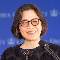 A photo of a woman with chin length dark hair who is wearing glasses and a dark shirt. She's speaking in front of a microphone.