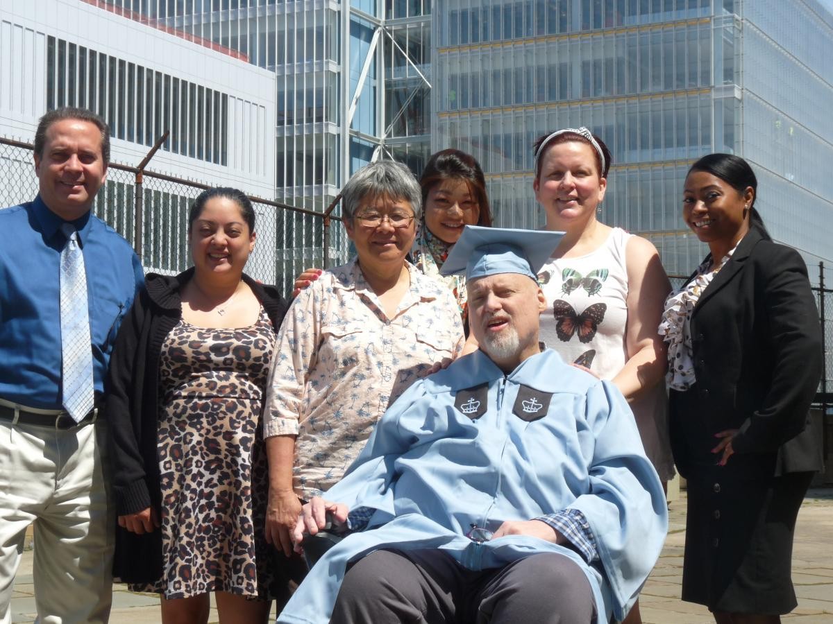 Edward Lauth (seated) surrounded by the friends, family and colleagues who helped him get through a degree program and breast cancer. Man seated in graduation cap and gown, surrounded by family