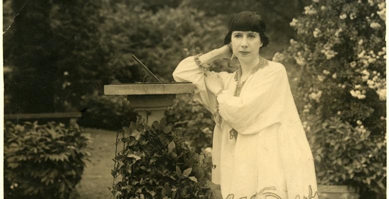 In a vintage black-and-white photo, a woman stands in a garden, leaning on a pedestal and wearing a flowing, white tunic