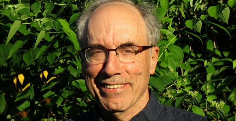 Leafy trees fill the background behind a man wearing glasses and smiling into the camera