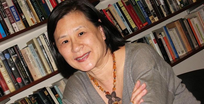 A woman, smiling slightly and wearing a gray V-neck sweater and a string of amber beads, sits in front of shelves lined with books