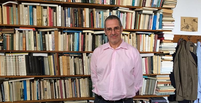 Mar Mazower in a pink button-down shirt while standing in front of a book shelf filled with books