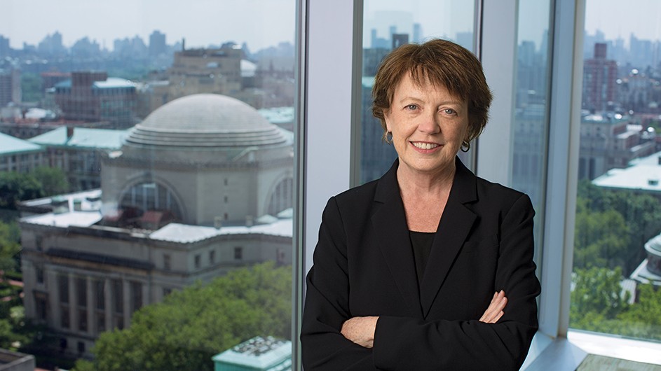 Mary Boyce stands in front of floor-to-ceiling windows with view over the Columbia University Morningside campus.