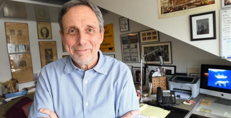 Prof. Michael Shapiro in his office at the journalism school. Photo by Eileen Barroso Wearing a blue dress shirt, with his arms folded across his chest, a man stand in an office, the walls of which are covered with framed photographs and news clippings