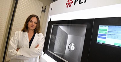 A woman wearing a white lab coat stands in front of a bank of lab equipment